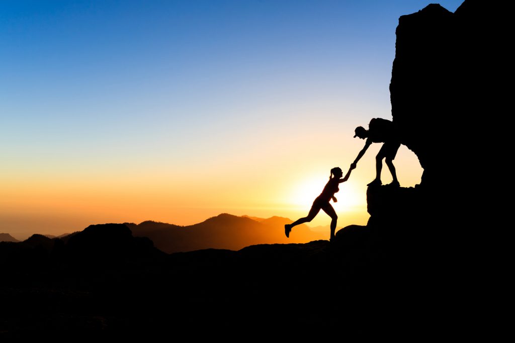 One partner helping another climb up a boulder against a sunset.
