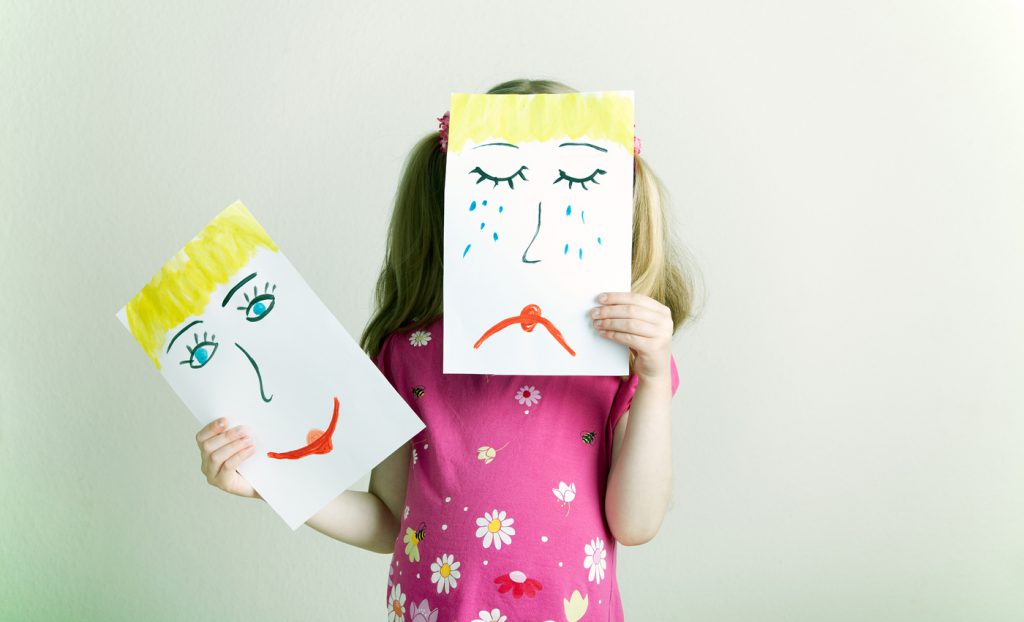 Little blonde girls holding happy and sad face masks symbolizing changing emotions