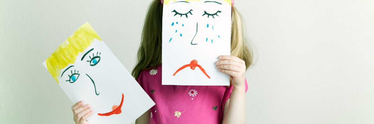 Little blonde girls holding happy and sad face masks symbolizing changing emotions