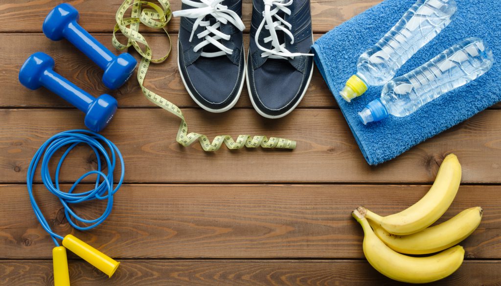 Fitness concept with sneakers dumbbells skipping rope measure tape towel bottle of water and bananas on wooden table background