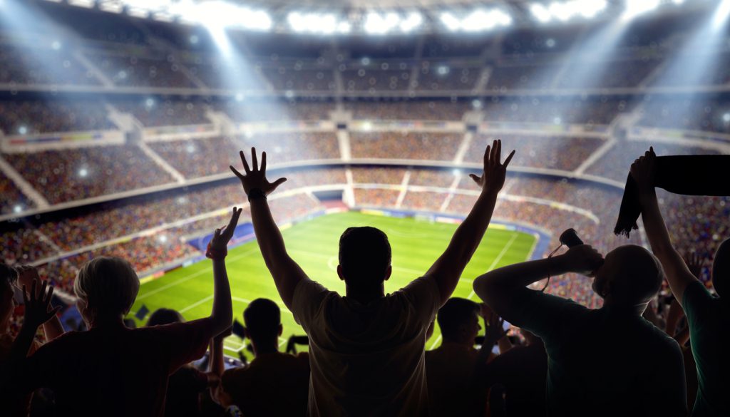 A long-range shot of a stadium field, floodlights and seating. A green field, with painted white lines, is visible in the foreground. On the foreground a group of fans is celebrating a goal. In the background are diffuse out-of-focus stadium seats. Large, bright floodlights are in the top-left and top-right corners of the image.