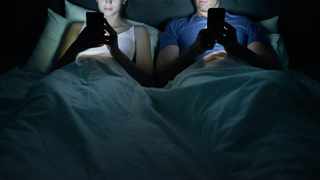 Cropped shot of a young couple using their cellphones in bed at night