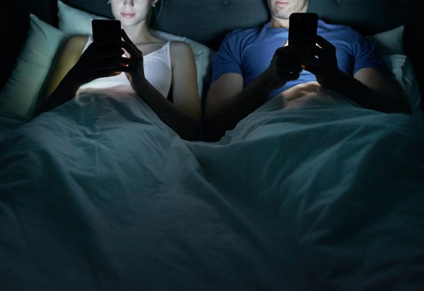 Cropped shot of a young couple using their cellphones in bed at night
