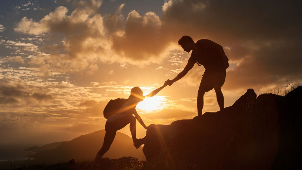 Male and female hikers climbing up mountain cliff and one of them giving helping hand.