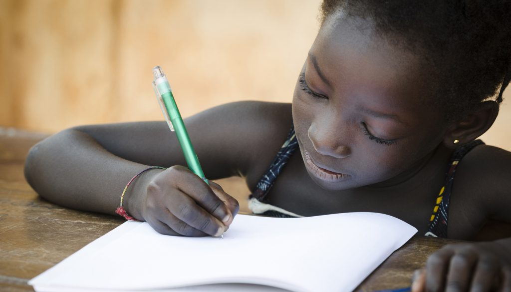 A young African girl writing her lesson on a sheet of paper.
