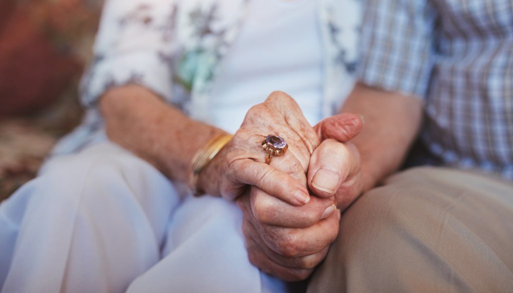 Cropped shot of elderly couple holding hands while sitting together at home.