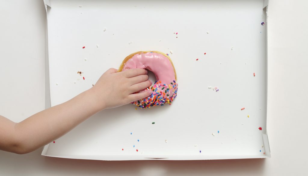 Overhead view of a doughnut box with a young girl's hand reaching to grab the last pink strawberry frosted donut.