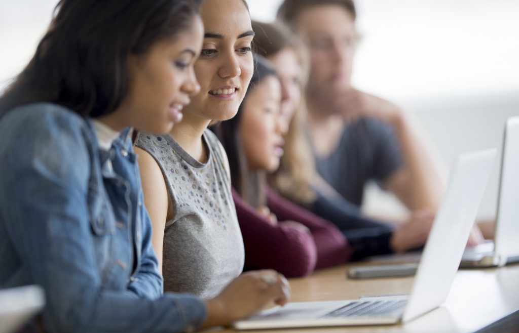 A multi-ethnic group of high school age students are sitting in class at are doing research on a laptop computer.