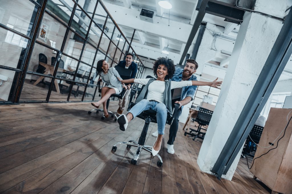 Four young cheerful business people in smart casual wear having fun while racing on office chairs and smiling