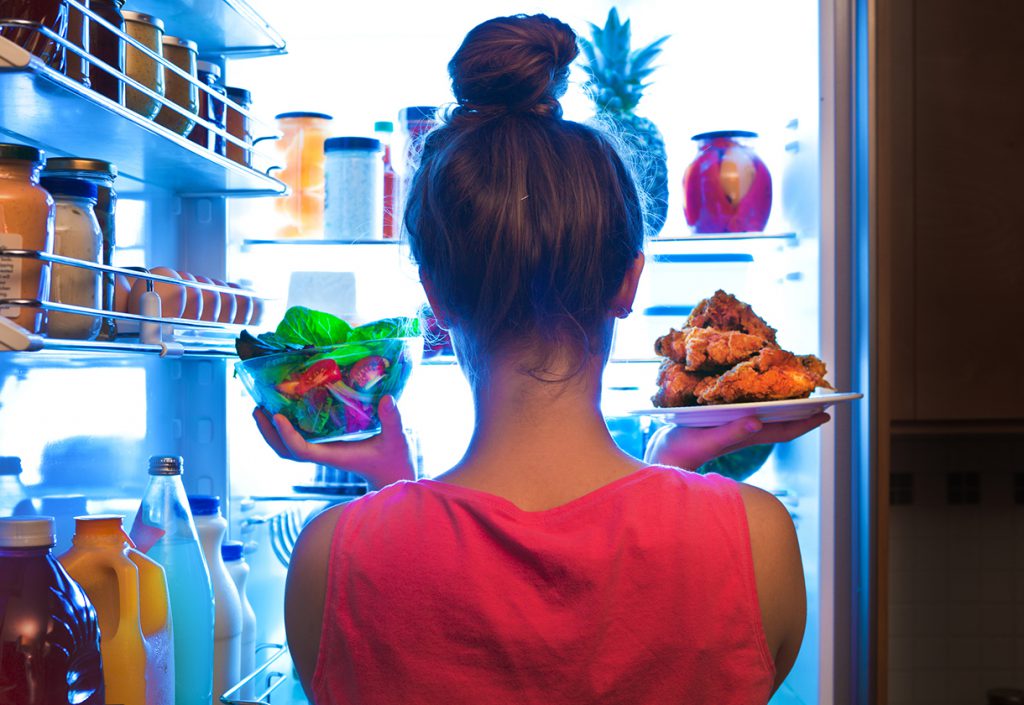 A young woman standing in front of the refrigerator, holding a bowl of salad in one hand and a plate of fried chicken in the other.