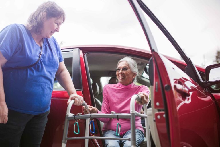 Woman helping another woman using a walker as she gets out of a car.
