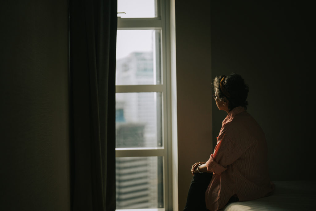 lonely senior woman standing in the dark looking out a window