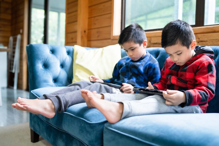 Two young boys sitting on a couch looking at tablets.