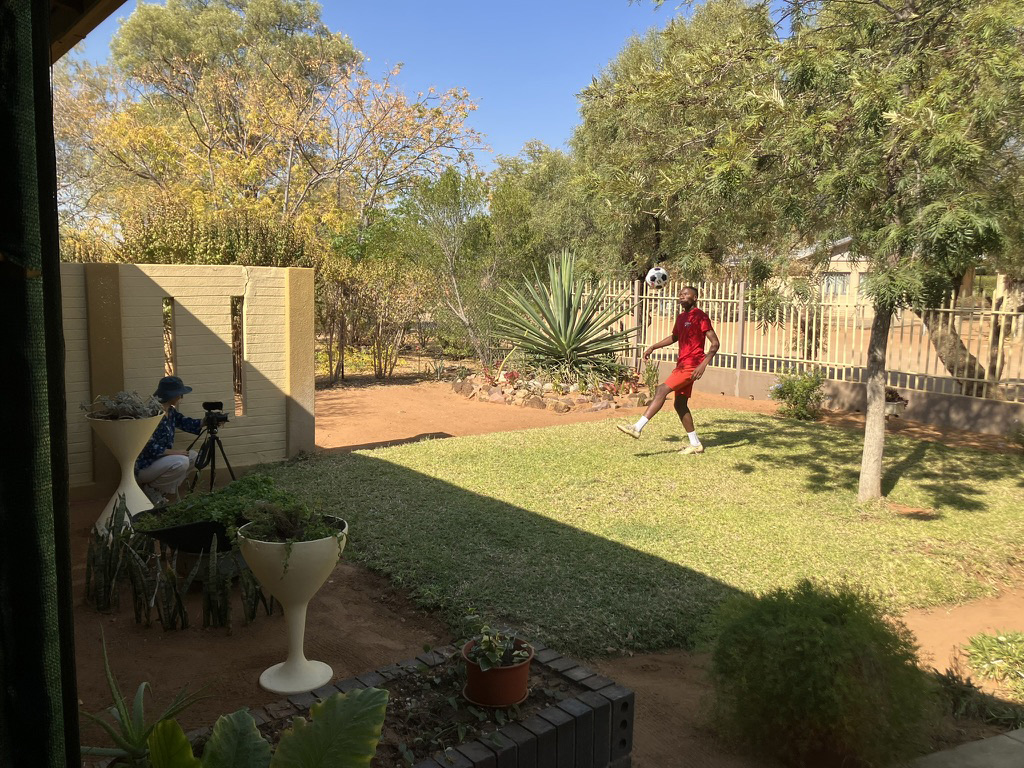 The young man plays soccer in a fenced in yard, with green foliage surrounding him.