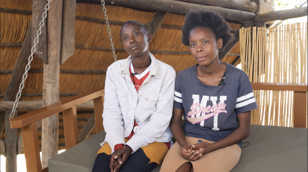 The women sit on a large outdoor swing, looking at the camera solemnly.