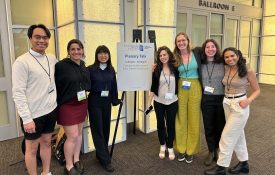 Six students and Julia Leonard pose for a photo around a sign regarding the plenary talk.