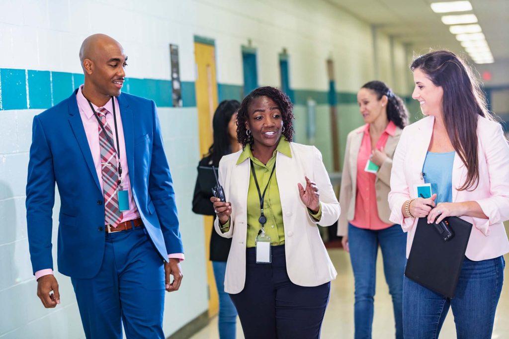 School teachers conversing as they walk through a school corridor.