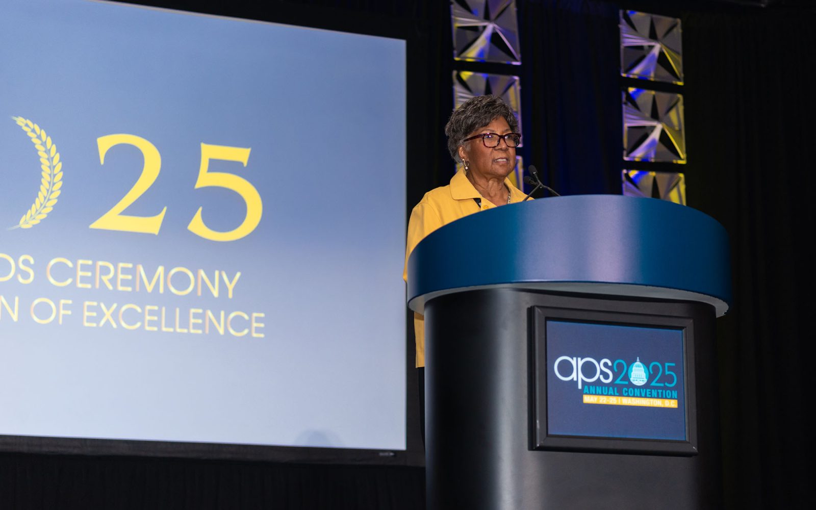 Margaret Beale Spencer stands at an APS 2025 branded podium as she gives her remarks.