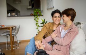 A lesbian couple sits on the couch together. They look and dress similarly.