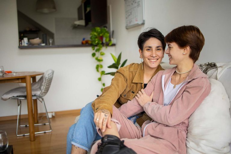 A lesbian couple sits on the couch together. They look and dress similarly.
