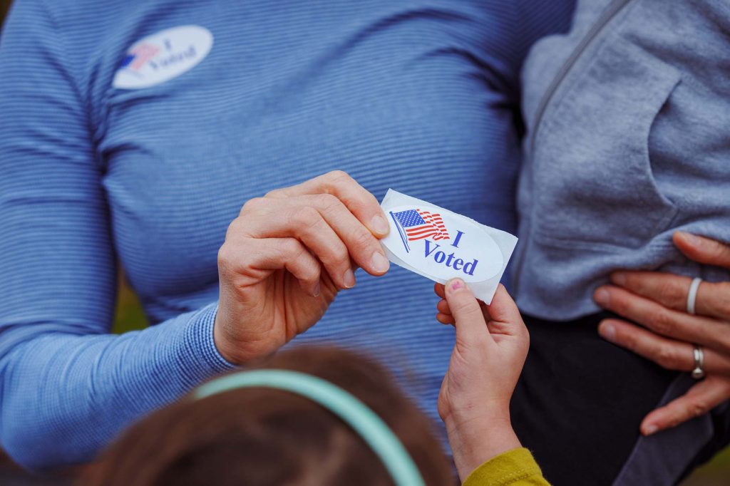 Cropped view of a woman holding her toddler on her hip and handing a sticker that says "I Voted" to another child.