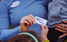 Cropped view of a woman holding her toddler on her hip and handing a sticker that says "I Voted" to another child.