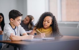 A multiracial middle school boy and girl sit at a classroom table while writing in open notebooks, solving schoolwork problems together.