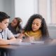 A multiracial middle school boy and girl sit at a classroom table while writing in open notebooks, solving schoolwork problems together.