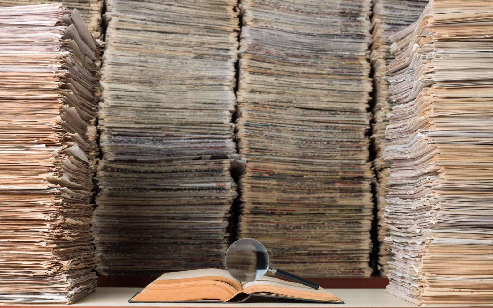 View of large amount of documents, newspapers, and books in classic library. A magnifying glass seen on top of an open book on desk.