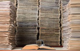 View of large amount of documents, newspapers, and books in classic library. A magnifying glass seen on top of an open book on desk.