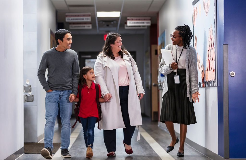 Two parents bringing their daughter to school, talking with an elementary school teacher or principal in the hallway.