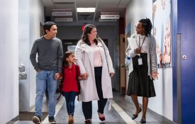 Two parents bringing their daughter to school, talking with an elementary school teacher or principal in the hallway.