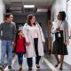 Two parents bringing their daughter to school, talking with an elementary school teacher or principal in the hallway.