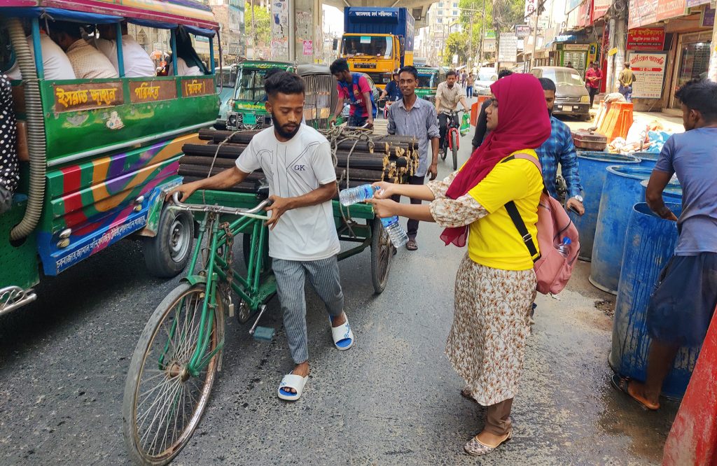A woman hands a man a water bottle as he walks his bike by.