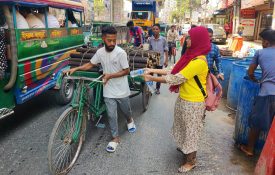 A woman hands a man a water bottle as he walks his bike by.
