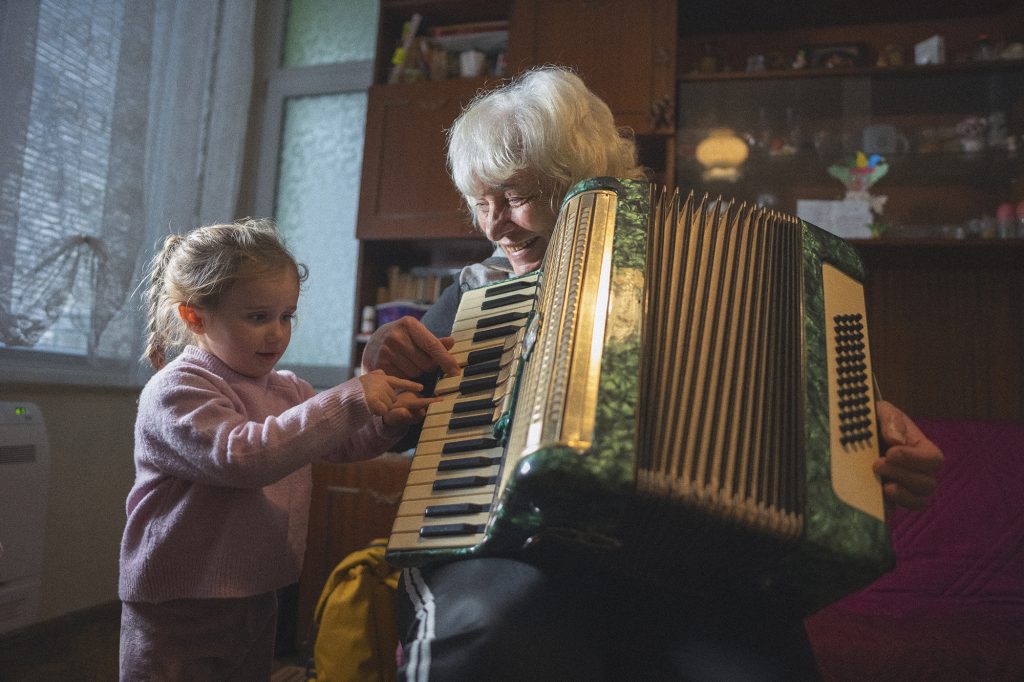 Multigenerational family moments at home. Grandma and granddaughter have fun while playing music on accordion together surrounded by soft light.