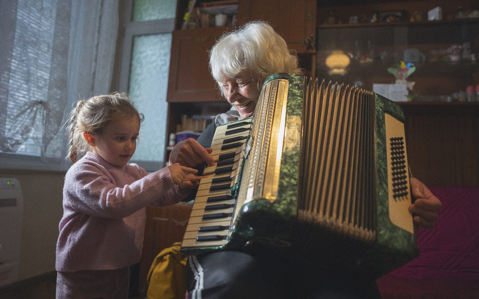 Multigenerational family moments at home. Grandma and granddaughter have fun while playing music on accordion together surrounded by soft light.