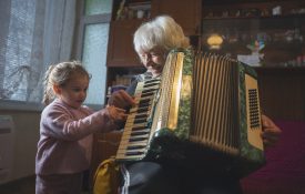 Multigenerational family moments at home. Grandma and granddaughter have fun while playing music on accordion together surrounded by soft light.