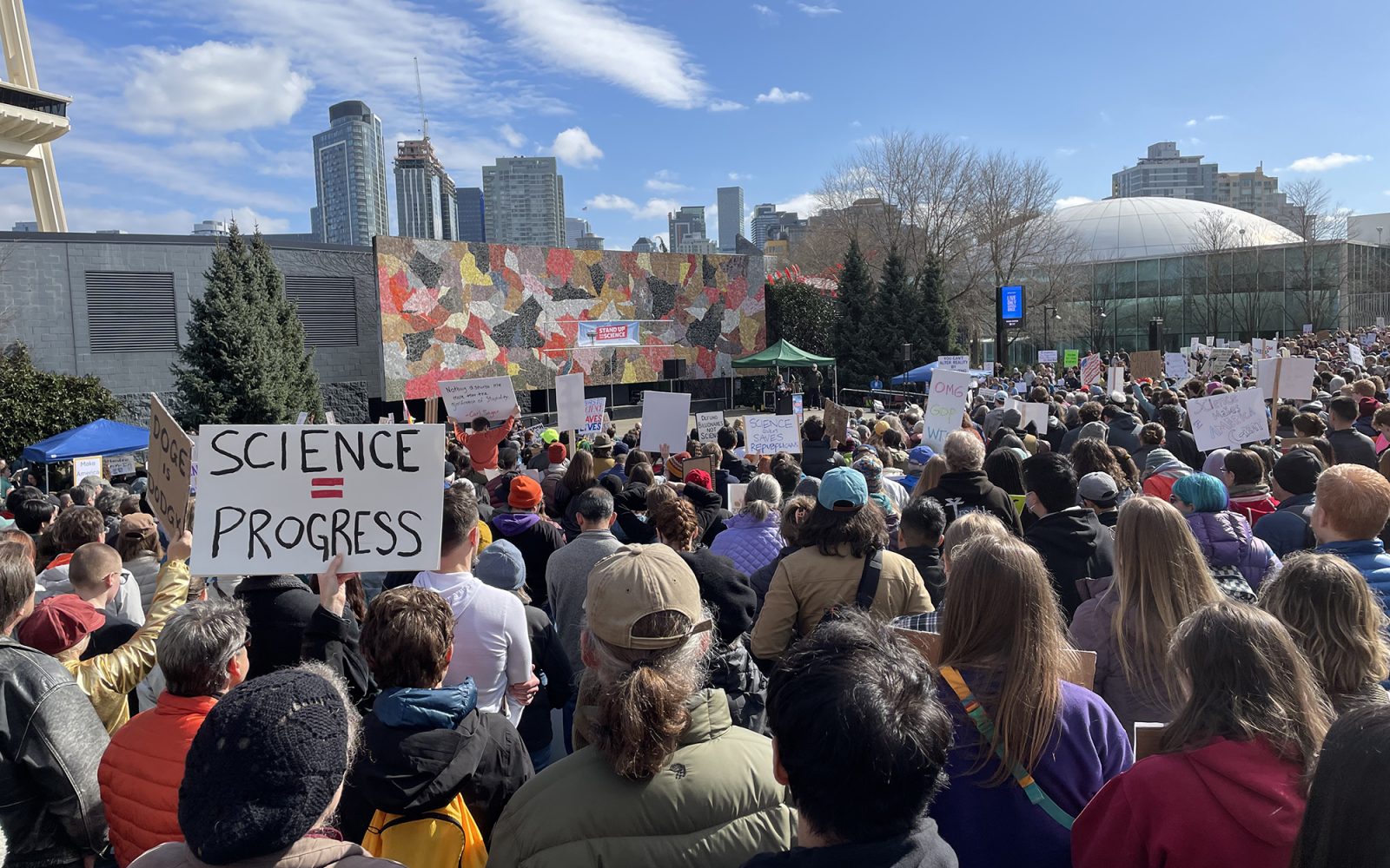 A photograph of a crowd of people at the Stand Up For Science protest in Seattle, Washington on 07 March 2025. Many people are holding signs, one of which says "Science = Progress." The people in the crowd are facing a speaker on a stage. At the top left of the photograph is part of the Space Needle, an iconic landmark in Seattle.