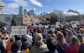 A photograph of a crowd of people at the Stand Up For Science protest in Seattle, Washington on 07 March 2025. Many people are holding signs, one of which says "Science = Progress." The people in the crowd are facing a speaker on a stage. At the top left of the photograph is part of the Space Needle, an iconic landmark in Seattle.