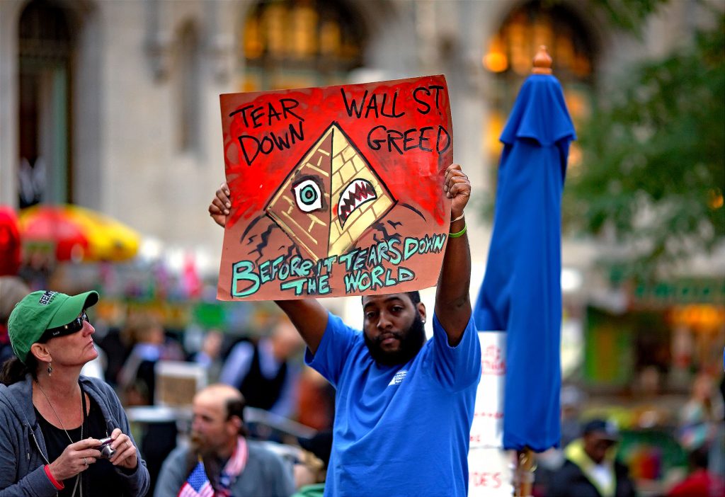 A Black man holding a protest sign during the Occupy Wall Street protest in 2011: "Tear down Wall St greed before it tears down the world."