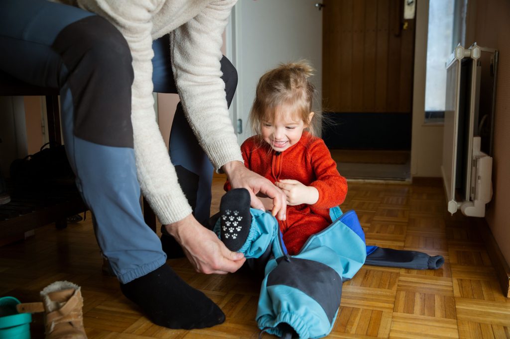 Father helping toddler get dressed in overall at hallway.