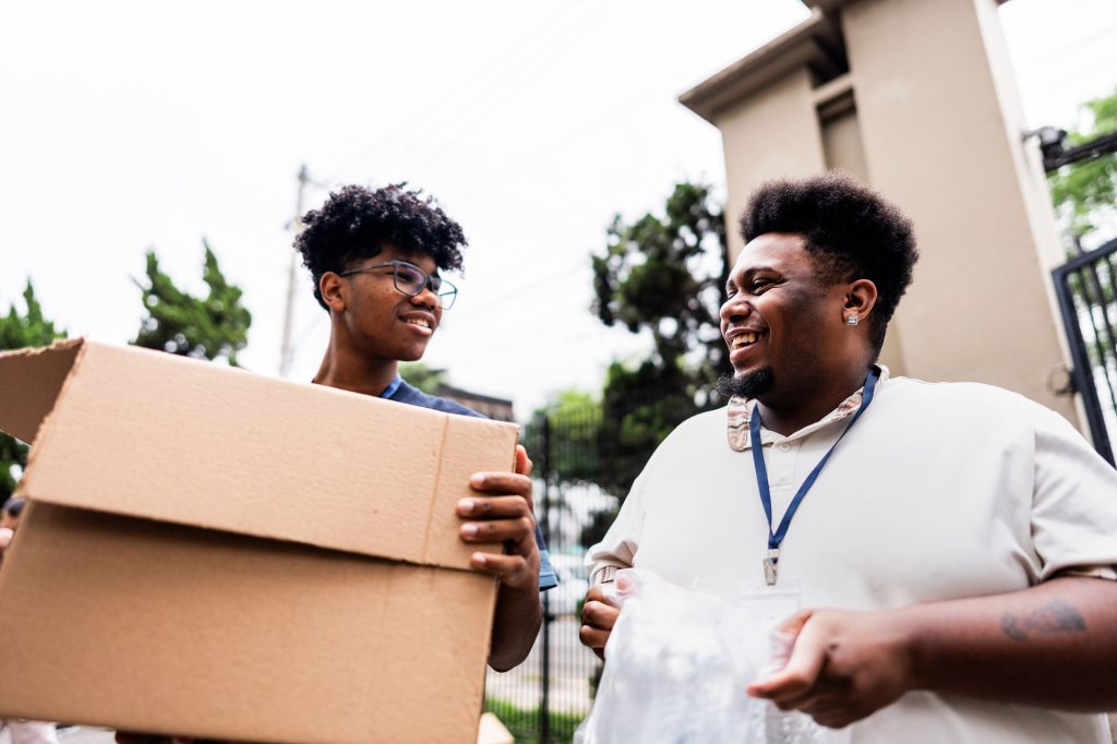 Two young volunteers work together during a community donation event, carrying cardboard boxes and communicating with enthusiasm.
