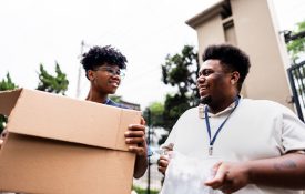 Two young volunteers work together during a community donation event, carrying cardboard boxes and communicating with enthusiasm.