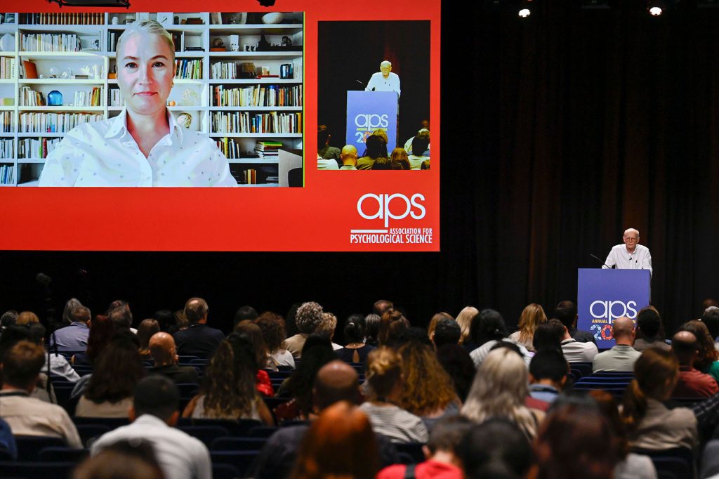 A packed plenary showing one virtual speaker, Rachael Jack, as she is being introduced by APS President James Pennebaker.
