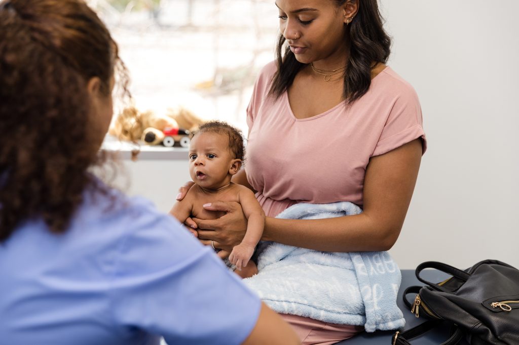Newborn boy sits in his mother's lap and attentively listens to a healthcare professional while she talks.