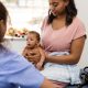 Newborn boy sits in his mother's lap and attentively listens to a healthcare professional while she talks.
