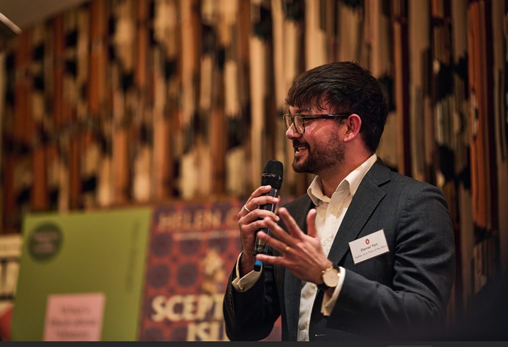 Yon holding a microphone and talking, with book covers behind him.