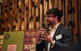 Yon holding a microphone and talking, with book covers behind him.