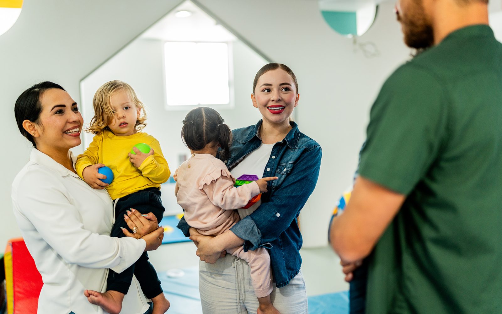 Parents carrying babies and talking at playroom.